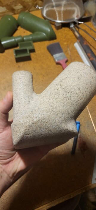 En hand håller ett nygjutet alujumjum-föremål i ljusgrå betongliknande material på ett arbetsbord.