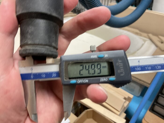 Hand med skjutmått mäter yttre diameter på en dammsugarslang, display visar 24,99 mm. Bakgrund visar träverktyg och en annan slang.