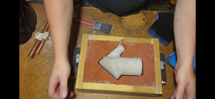 Gjuten pilform i sandform, träkantad ram, pensel och kopparrör på arbetsbord.