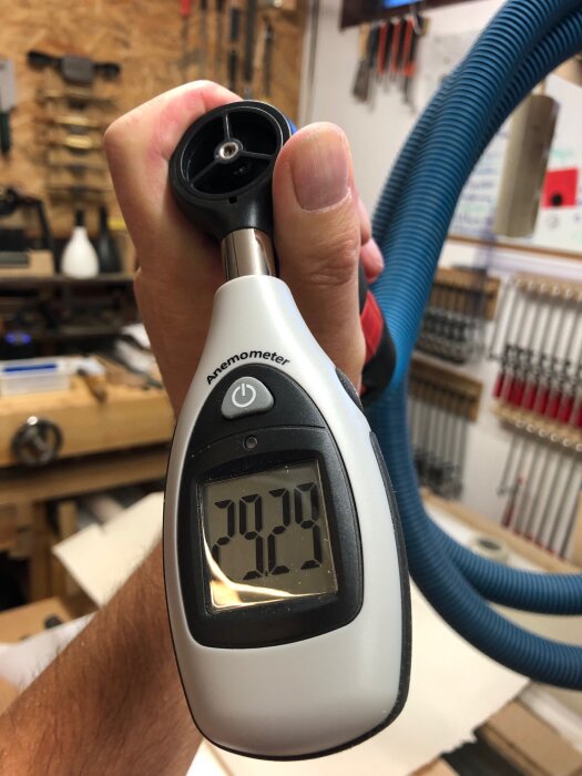 Hand håller en anemometer som visar 29,29 m/s, med en blå dammsugarslang i bakgrunden i verkstadsmiljö.
