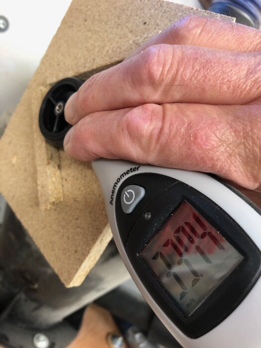 Hand håller anemometer med display som visar hastighet 33,94 m/s, mätning av luftflöde genom hål i träplatta.
