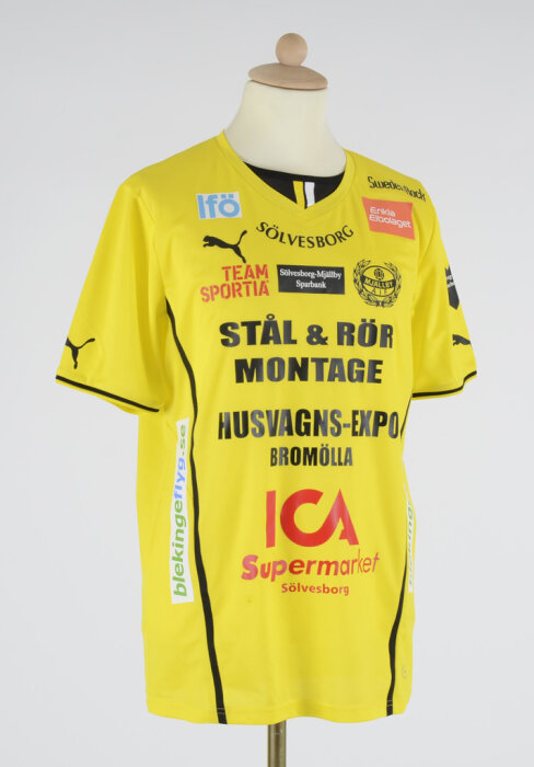 En gul fotbollströja med olika sponsorer som ICA, Team Sportia och Husvagns-Expo Bromölla tryckta på framsidan.