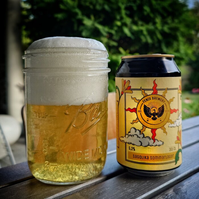 Ölglas med skummande lager bredvid en burk Fenix Brewery Sagolika Sommarlager på ett bord utomhus.