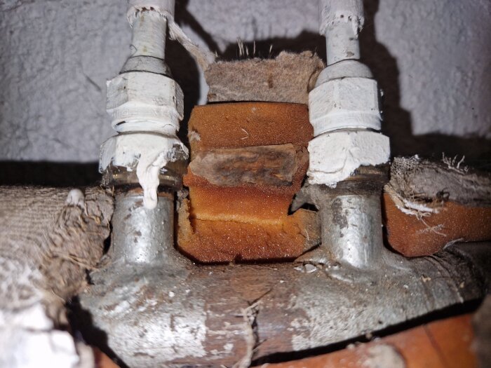 Två metallrör med vita kopplingar, omgivna av isolering, anslutna till ett större huvudrör.