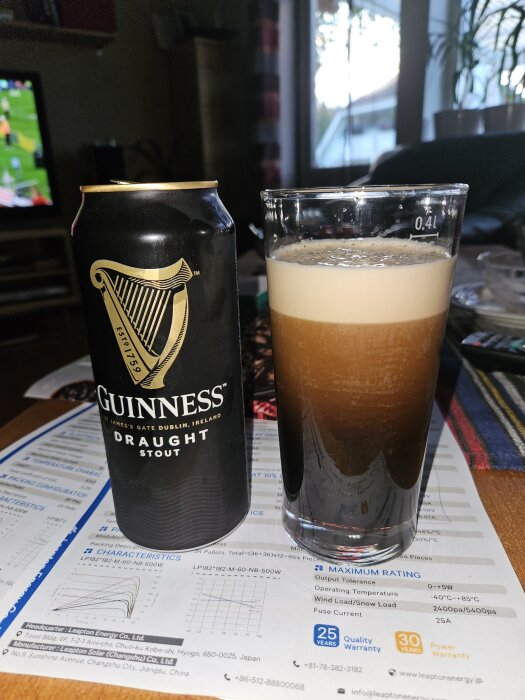 En burk Guinness och ett glas med skummande öl står på ett papper med diagram, i ett vardagsrum med TV i bakgrunden.