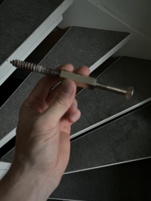 Hand håller en stor skruv med distans ovanför trappsteg, redo för montering av en ledstång.