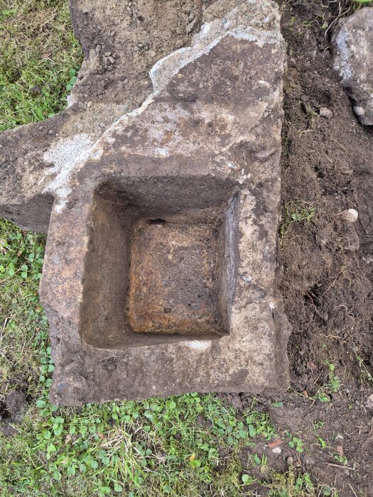 En grävd betongkonstruktion med en fyrkantig urgröpning och ingjutet järnrör längst ner, omgiven av gräs och jord.