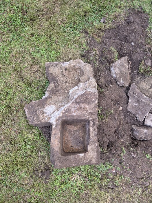 Betongkonstruktion i gräsmattan, delvis uppgrävd. Fyrkantig hålighet synlig, omgiven av gräs och jord. Eventuellt inslag av järnrör.
