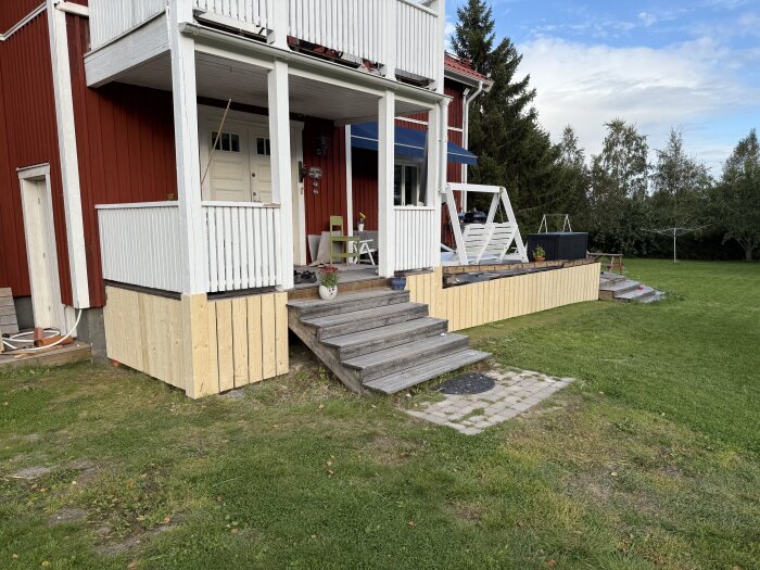 Nybyggd farstukvist och altan med omålade bräder, sittgrupp och klätternät till barnens trädkoja, omgivna av grön gräsmatta och träd.