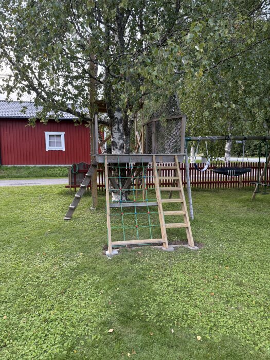 Klätternät och trappa till barnens trädkoja placerad vid ett träd, omgiven av grönska och med en röd stuga i bakgrunden.