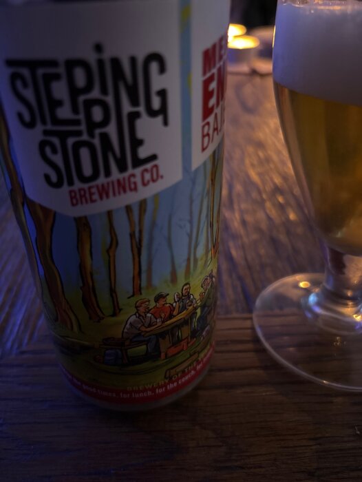 Burk med Stepping Stone Brewing Co. och Mere End Bajer etikett bredvid ett glas öl på ett bord i dämpad belysning.