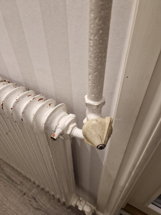 En närbild av ett vitmålat, äldre element med en ansluten rörledning och en ventil med plasthandtag.