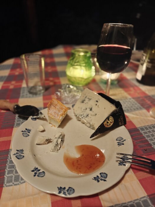 Osttallrik med olika ostar och marmelad pÄ en blommig tallrik, glas rött vin och ljuslykta i bakgrunden pÄ en rutig duk.