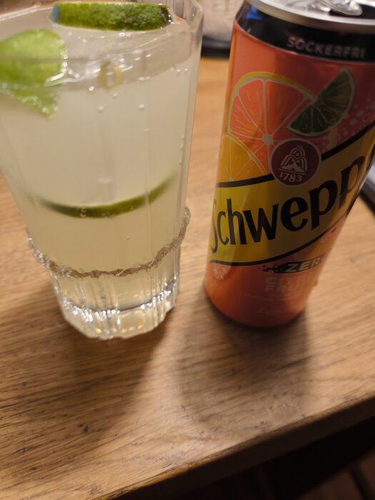 En drink med Absolut Vodka, Schweppes Citrus och lime i ett glas bredvid en Schweppes burk på ett träbord.