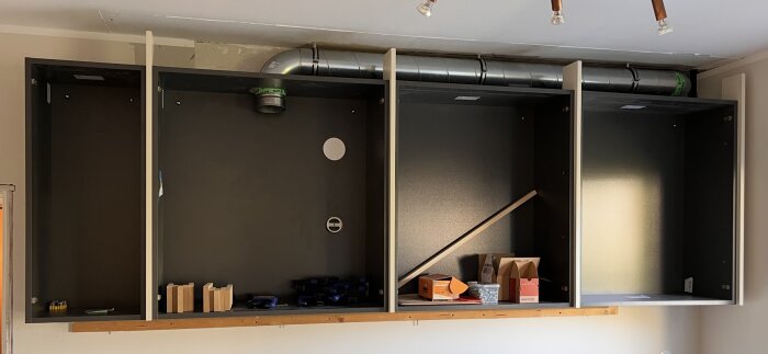 Överskåp monterade på vägg med synlig ventilation ovanpå, mellanlist delvis monterad. Verktyg och material i skåpen.