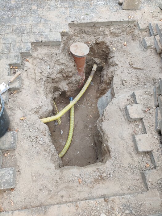 Grop framför entré med synliga rör och jord, där ett gult skyddsrör ligger i marken. Murstenar och verktyg syns vid sidan.