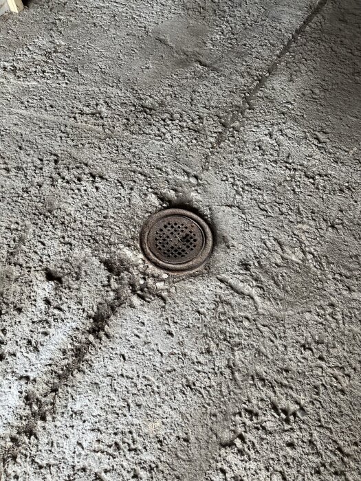 Grovt betonggolv i garage med ett runt metallbrunnslock, ytan är ojämn och i behov av flytspackling.
