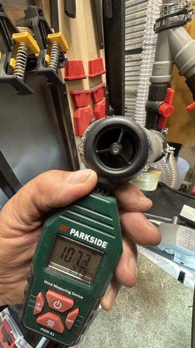 En hand håller en Parkside-anemometer som visar 107,3 km/h bredvid ett cyklonsystem med rör och slangar i en verkstadsmiljö.