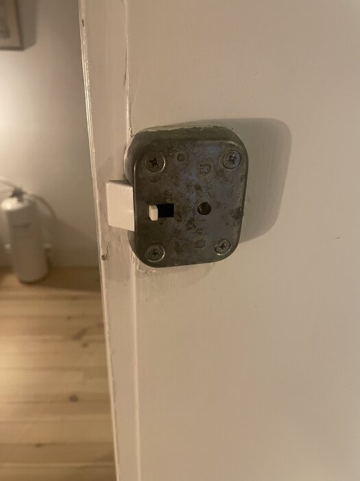 Gammalt dörrlås med bristfälligt insideshandtag, monterat på en vit dörr, där insidan visar fyra skruvar och ett befintligt nyckelhål.