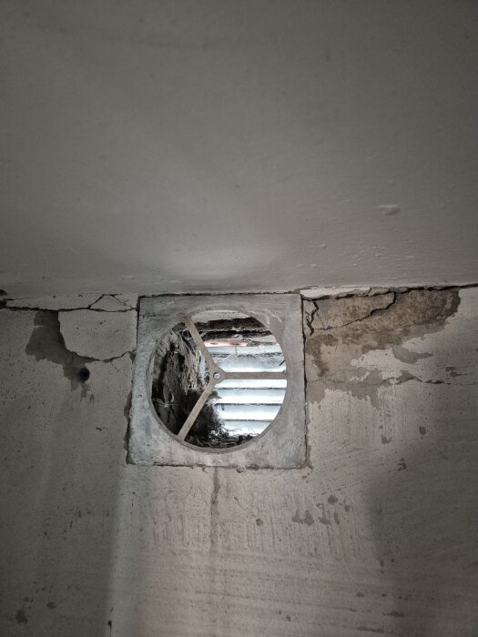 En igensatt ventil i en källarvägg, omgiven av sprucken puts och fuktfläckar.