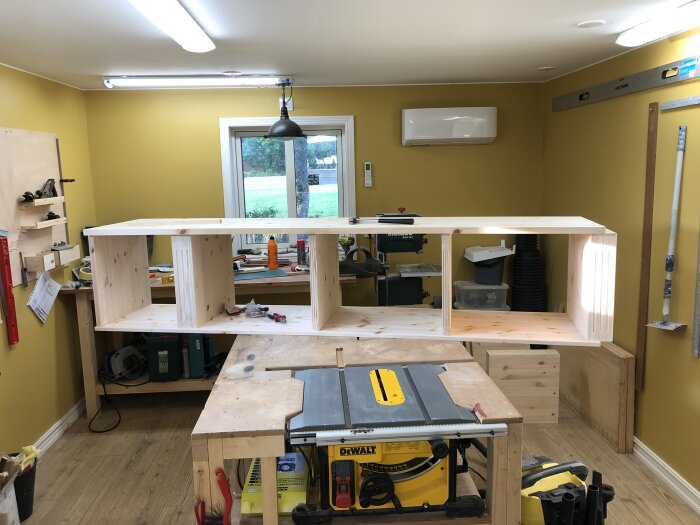 Trähylla under konstruktion i verkstad med furuplankor, arbetsbänk och verktyg, belyst av taklampor, mot en gul vägg.