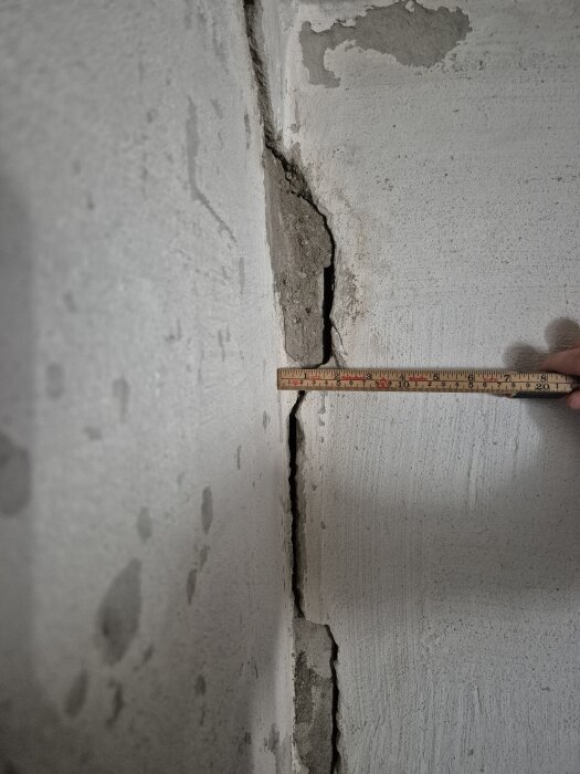 Spricka i betongvägg med tydliga lager av puts och synlig mätsticka för skala. Bakgrund av ojämnt målad vit yta.