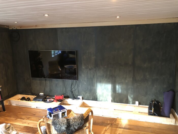 Bredd vägg med monterad TV över ett bygge av en hylla med inbyggd sockel, trädetaljer och elektriska enheter, i ett rum med trägolv och takpanel.