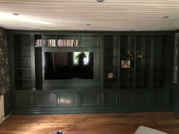 Grönmålad platsbyggd hylla med TV i mitten, omgiven av förvaringshyllor och skåp i trä, dekorerad med böcker och prydnader.