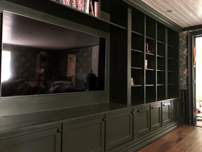 Grönmålad inbyggd hylla med öppna sektioner och skåp, integrerad TV och detaljer i klassisk stil. Blommönstrad vägg i bakgrunden.