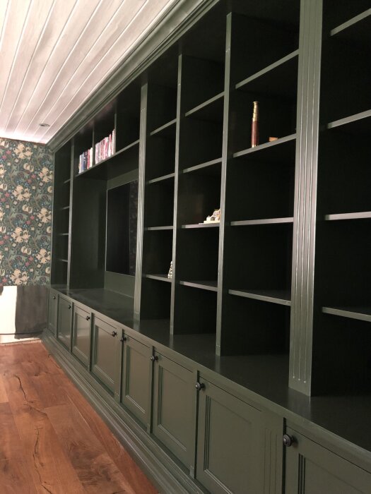 Grön måttbygghylla med TV-utrymme och hyllor, gjord av furu och ply, mot mönstrad tapet.