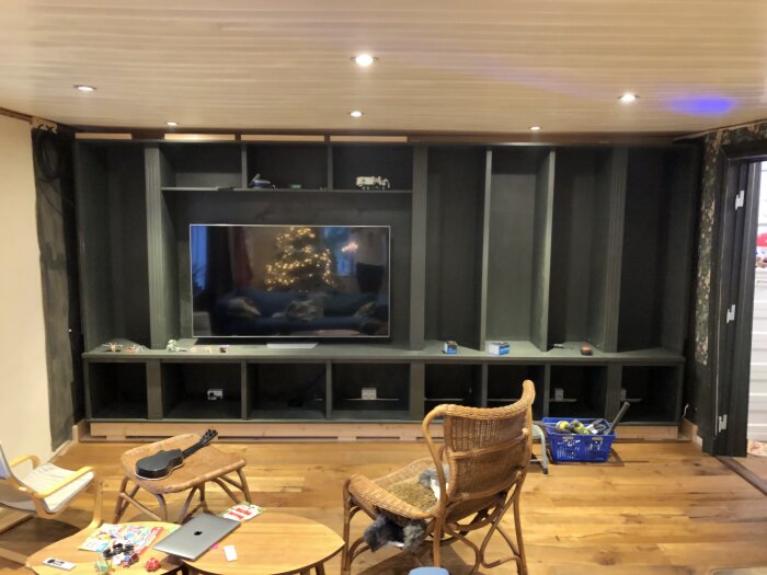 Grön, inbyggd hylla med 65" TV, förvaring och hyllor i ett rum med trägolv och rottingmöbler. Julgran synlig i TV-skärmen.