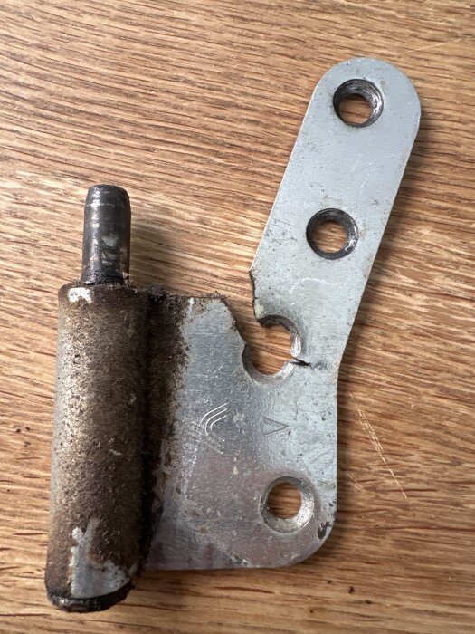 Använd ytterdörrens trasiga nedre gångjärn med skruvhål, tydligt slitet och delvis rostigt, liggande på en trägolv.