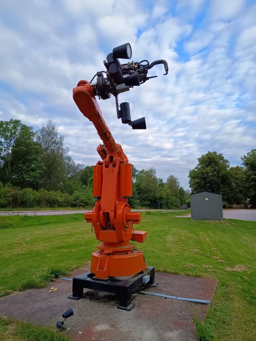 Orange industrirobot som nu är en installation vid Sverob i Skogstorp, omgiven av grönska och en asfalterad väg i bakgrunden.