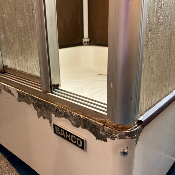 Föråldrad duschkabin med rostiga och slitna metalldelar samt Bahco-märke, relaterat till diskussion om renoveringsbehov i äldre badrum.