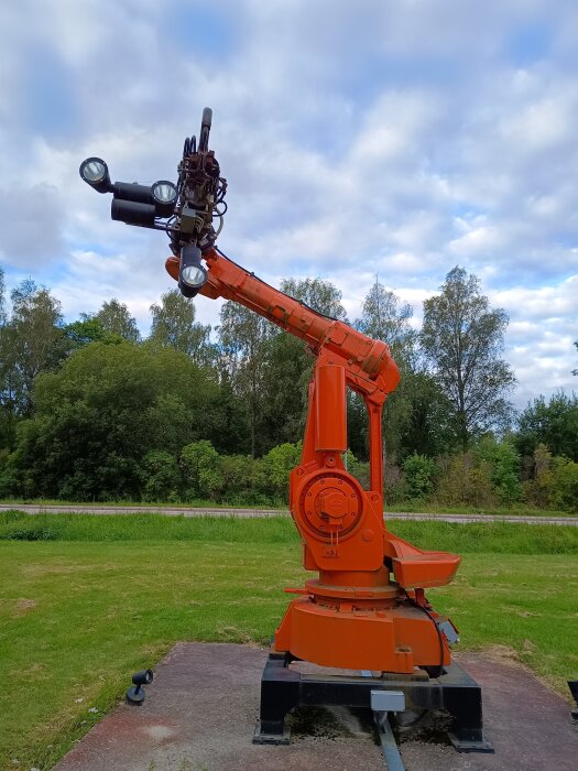 Orange industrirobot på en platta utomhus, omgiven av grönska, tidigare använd i industrin, nu utställd vid Sverob i Skogstorp.