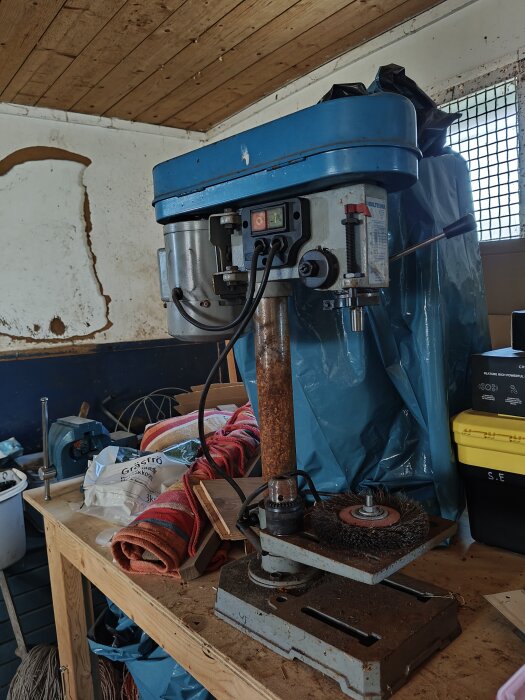 Äldre bänkpelarborrmaskin av märket Biltema med rost och slitage, placerad på ett arbetsbord i ett verkstadsutrymme.