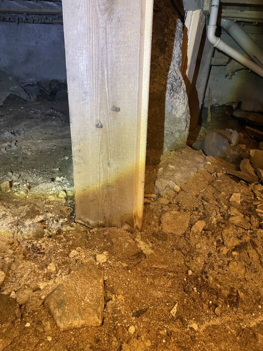 Träregel och stenfundament i en krypgrund under renovering, med jord och rör i bakgrunden.
