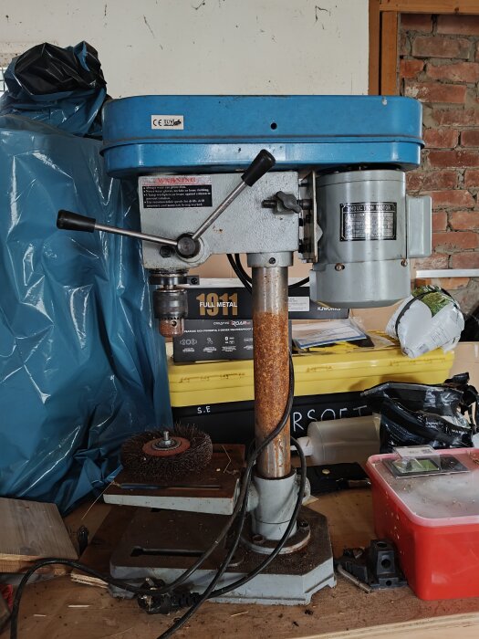 Äldre bänkpelarborrmaskin från Biltema med ytrost, sett på en arbetsbänk omgiven av verktyg och förpackningar.