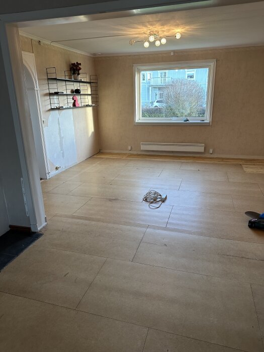 Rum under renovering med ett synligt undergolv, sladdar på golvet och ljus från fönstret.