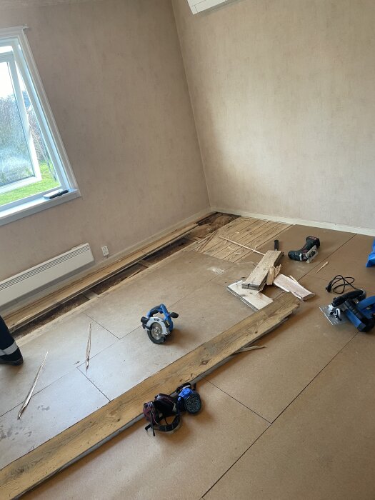 Ett rum där golvet har rivits upp, verktyg och träbitar ligger spridda. Cirkelsåg och hörselskydd syns på plywoodgolvet.