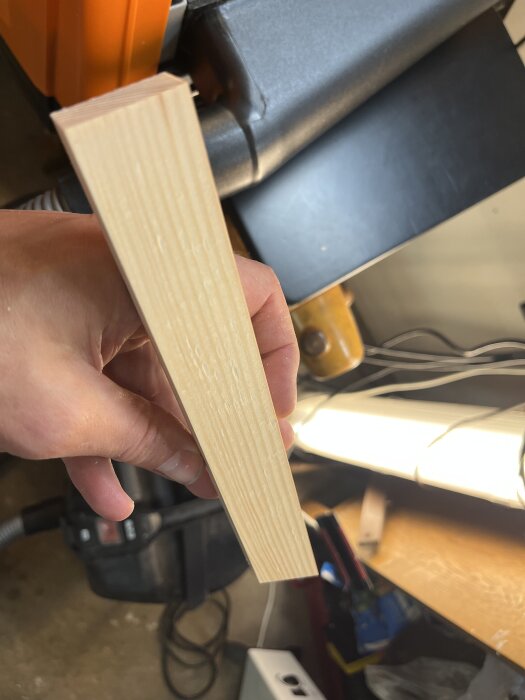 Hand håller en bit bearbetat furuträ framför en verkstadsmaskin.