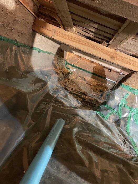 Plastfolie utspridd över en grund i en källare för att skydda mot fukt, med en blå rörsektion och uppbyggda träreglar ovanför.