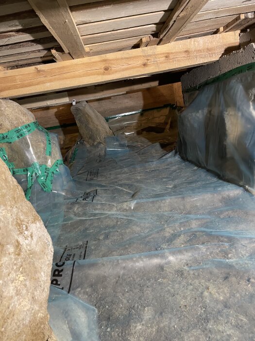 Plastfolie lagd över jordgolv i krypgrund under en träkonstruktion, med gröna tejpfogar och stora stenar synliga på sidorna.