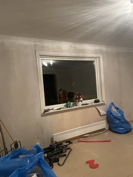 Rum under renovering med spacklade väggar, ett stort fönster och verktyg på golvet. Blå sopsäckar till höger.
