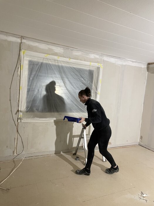 En person i arbetskläder gör förberedelser inför målning i renoveringsprojekt, plast över ett fönster, med spackelverktyg i handen.