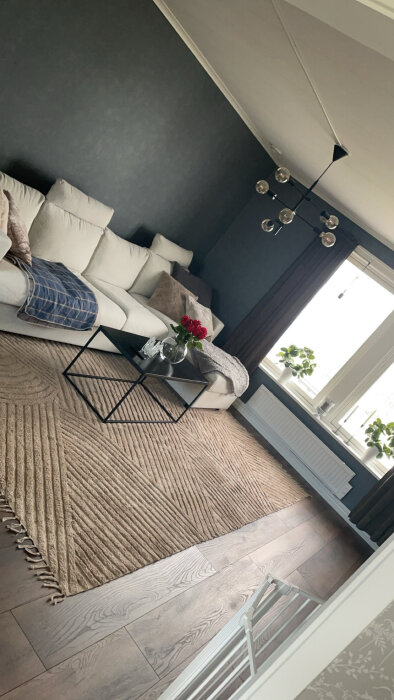 Ljust vardagsrum med grå vägg, vit soffa, mörk soffbord, beige matta och modern lampa. Fönster med gröna växter och vita persienner.