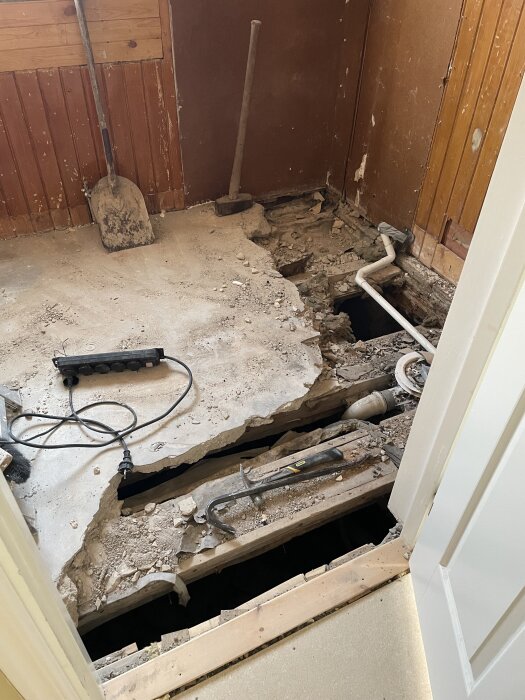 Renovering av toalett/entré med trasigt golv, blottade rör och verktyg; spade och slägga mot väggen.