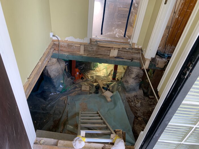 Renovering av hall med borttaget golv och synlig bärlina, stege leder ner till utrymme fyllt med plast och byggmaterial.