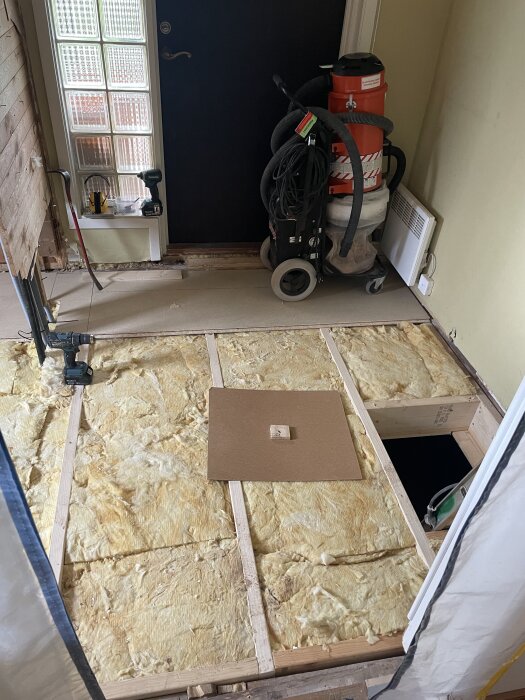 Renovering pågår: isolering läggs, verktyg vid sidan, borrmaskin, och en industridammsugare står redo. Trägolv under konstruktion.