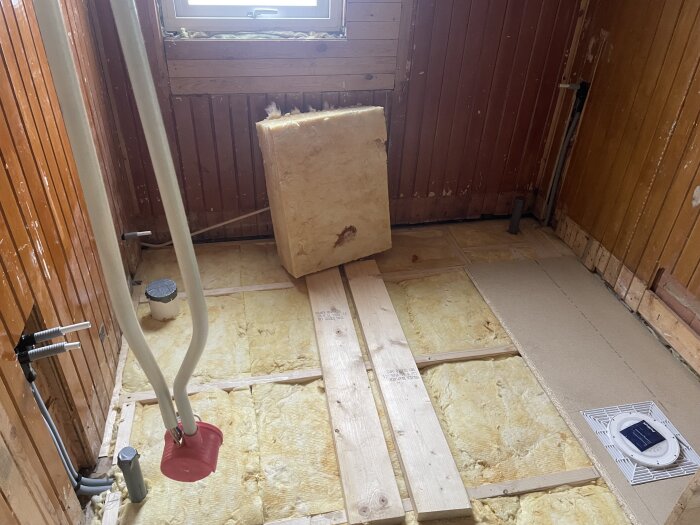 Rum under renovering med nya rör, träbalkar, isolering, och en ventilationstrumma på golvet. Bruna träväggar omger rummet.